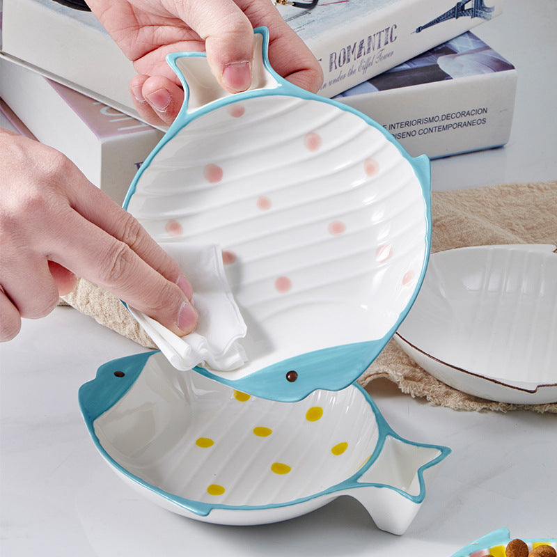 Hands wiping a pink polka-dot fish-shaped ceramic cat bowl, demonstrating its easy-to-clean dishwasher-safe glaze